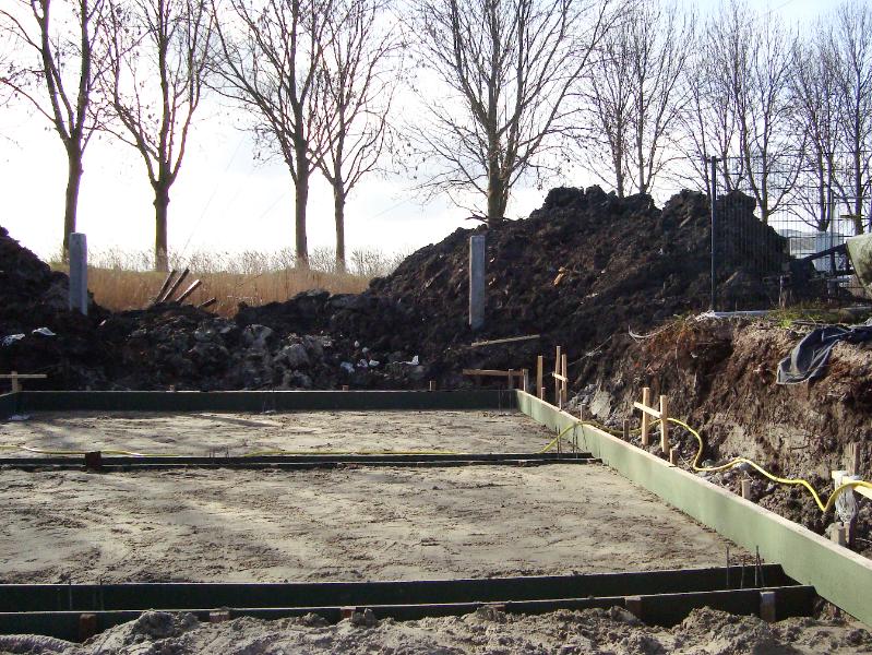 woodenhouse-013.jpg - De bekisting van de draagbalken en de randen van de betonvloer worden  uit goedkoop spaanplaat gemaakt Zie de vervuilde grond en de heipalen op de achtergrond die de hoogte van de begane grond markeren. Ook zijn de piket paaltjes te zien die de exacte plaats van het huis markeren. Het is belangrijk dat de muren exact 90 graden zijn.